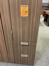 Load image into Gallery viewer, A tall wooden storage cabinet or wardrobe with a woodgrain finish, shown in a showroom. The cabinet has two doors on the upper left and two drawers on the lower right, with silver metal handles.