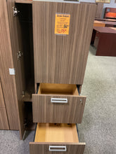 Load image into Gallery viewer, A tall wood-grain storage cabinet with two pull-out drawers, shown in a furniture store setting with carpet and other furniture visible in the background. A yellow price sticker is attached to the upper door.